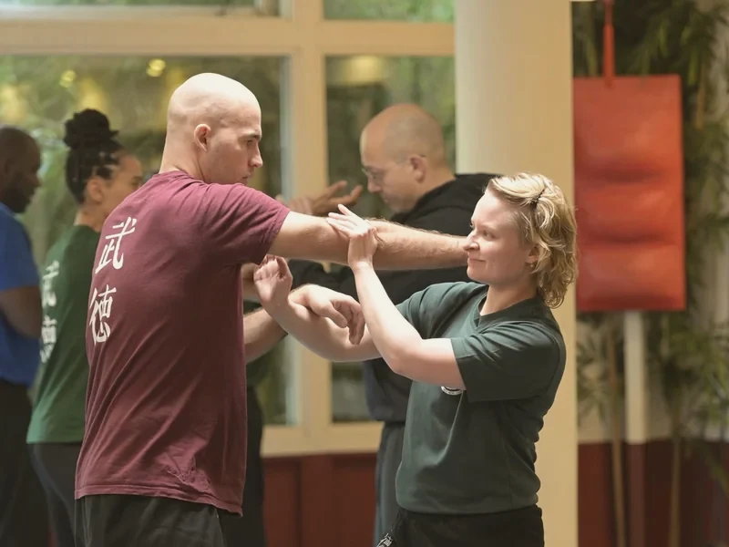 Twee leden oefenen samen een Wing Chun-armtechniek in de zaal; op de achtergrond trainen anderen